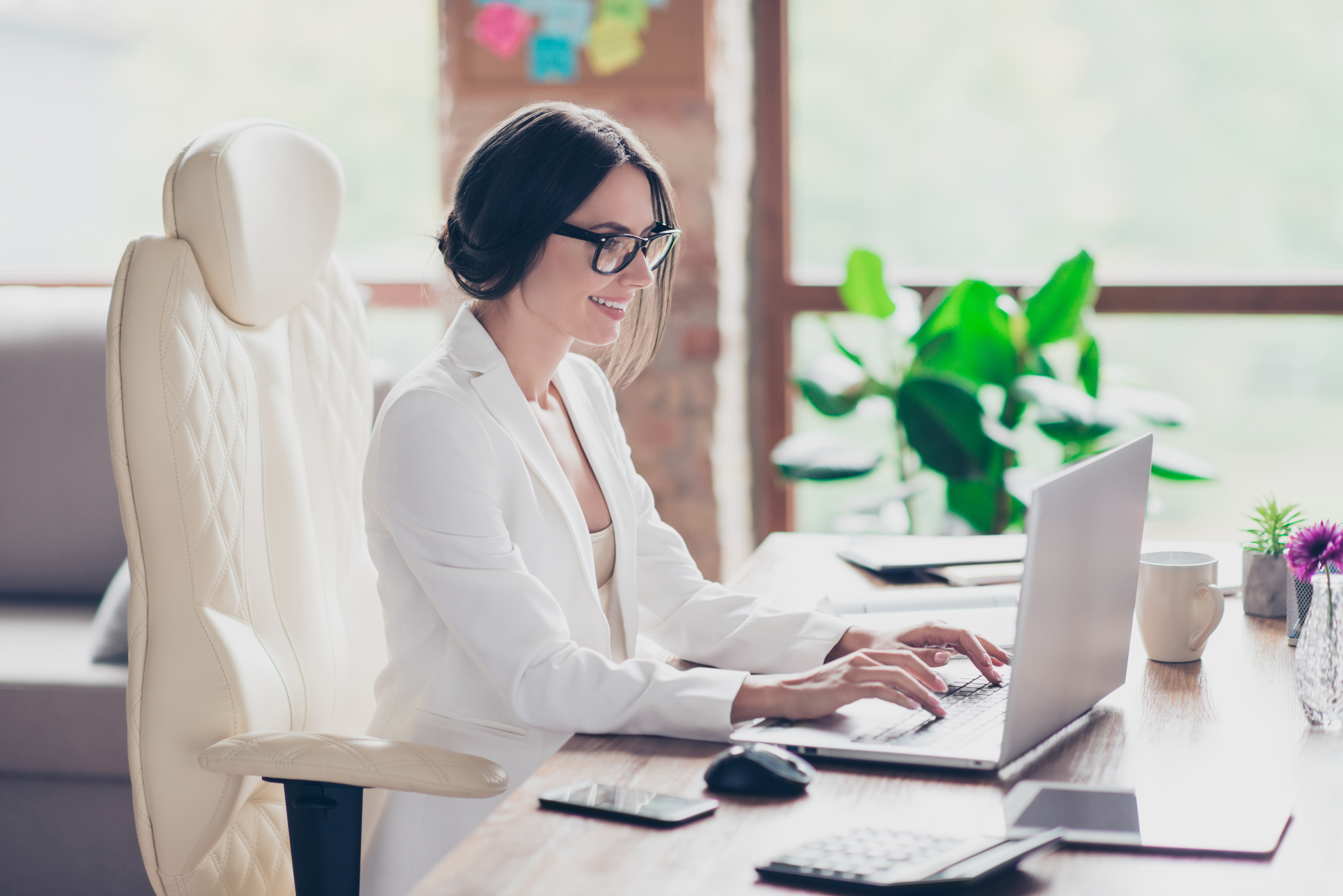 A smiling woman working on a laptop in a modern office, appearing focused and productive.