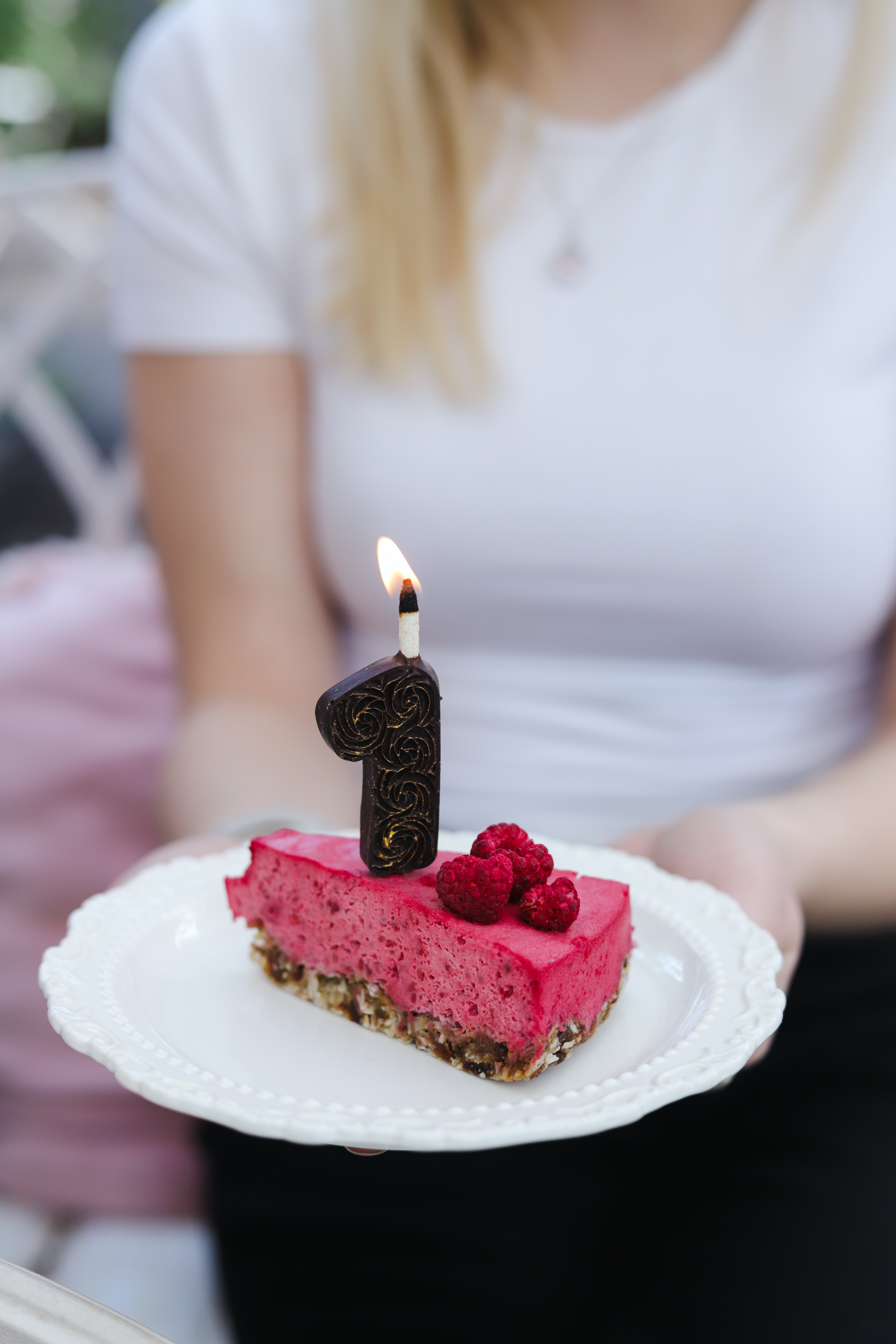 A slice of pink cake sits on a white plate with a lit number one birthday candle on top.