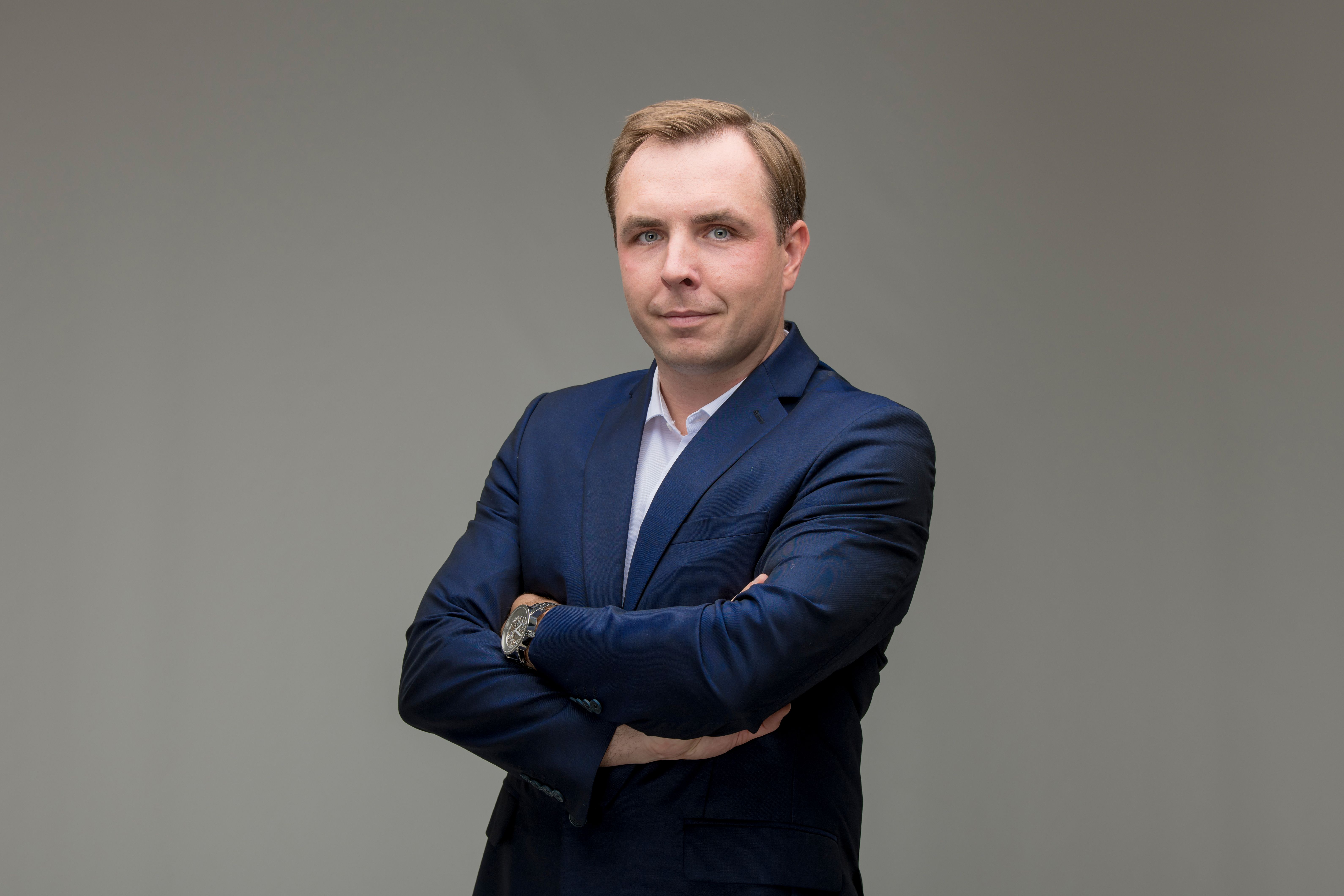 Mees tumesinises ülikonnas hallil taustal, käed rinnal.
