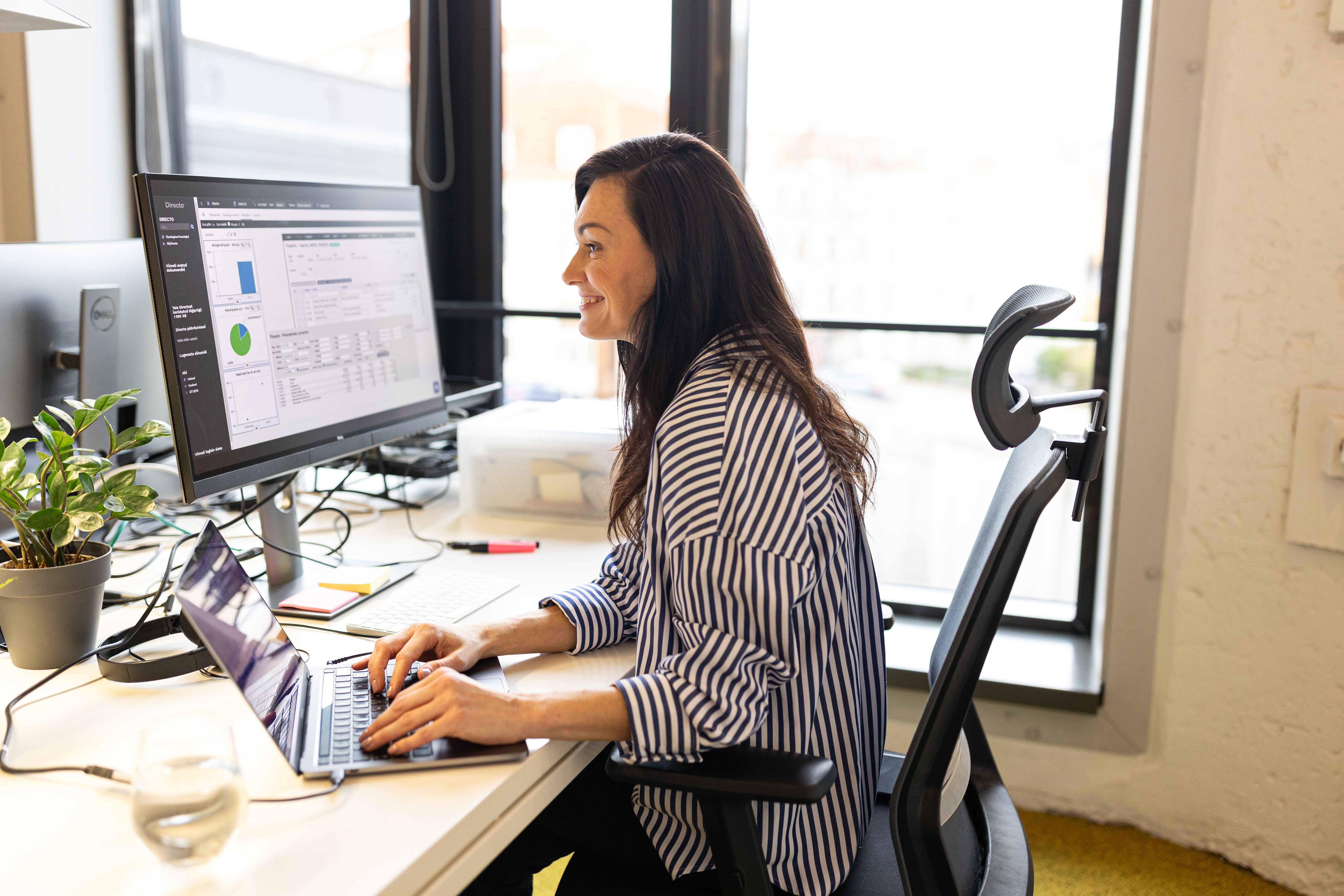 Naine töötab avatud kontoris ettevõtte loomise kallal. Arvutiekraanil on finants- ja ärianalüüs, naine naeratab ning tegutseb keskendunult.