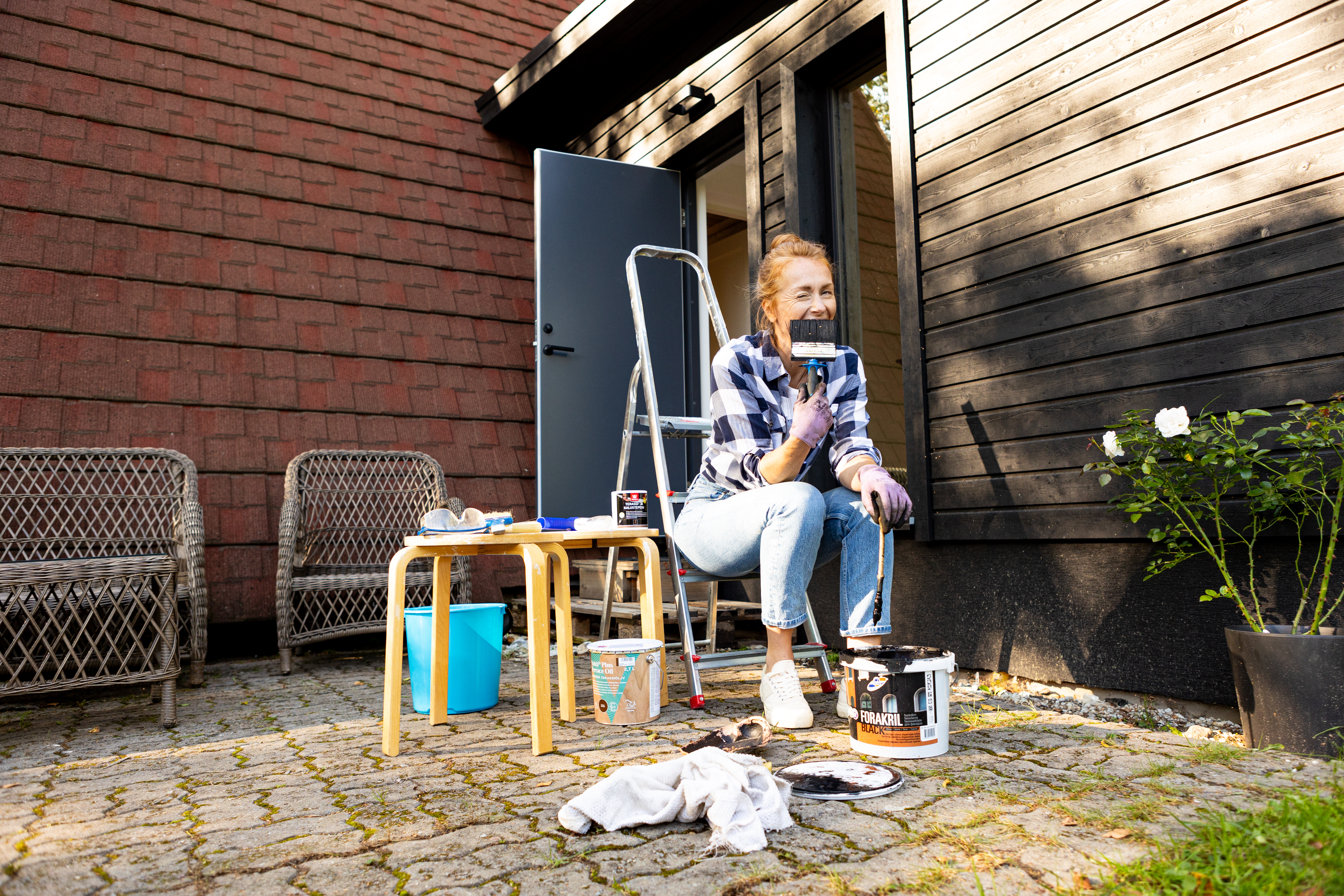 Naine värvib maja välisseina ja istub redelil värvipurkide kõrval.