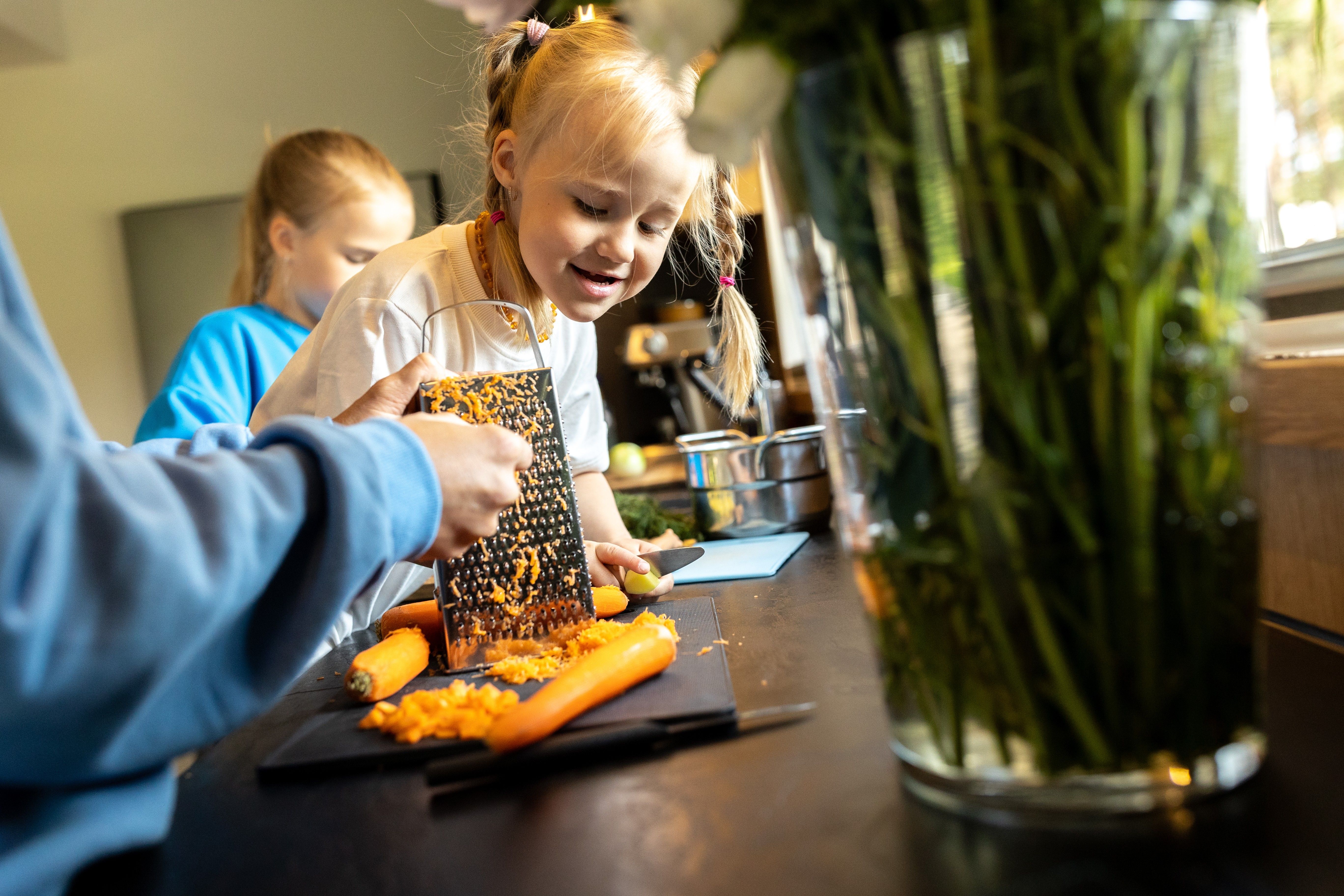 Lapsed valmistavad köögis koos toitu. Üks laps riivib porgandit, teine aitab tööpinna juures.